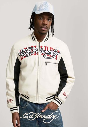 Młody mężczyzna z zaplecionymi włosami nosi białą i czarną kurtkę varsity z logo "Ed Hardy", niebieskie dżinsy z pasującym logo oraz jasnoniebieską czapkę.