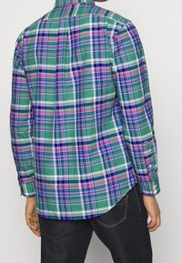 Green plaid shirt showcasing a mix of purple, white, and blue stripes, with long sleeves, a button-down collar, and a rounded hem.