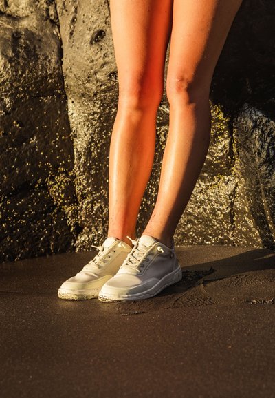 Baskets de sport blanches avec une tige en maille texturée, des lacets et des semelles en caoutchouc, posées sur du sable mouillé près d'une surface rocheuse.