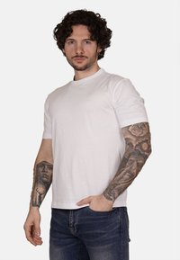 T-shirt bianco in cotone con design a girocollo. Maniche corte e un logo ricamato discreto sul petto. Vestibilità casual. Tatuaggi color pelle sulle braccia.