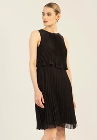 Schwarzes plissiertes Kleid mit ärmellosem Design, fließendem Stoff und mehrlagigem Saum. Es hat einen hohen Ausschnitt und eine weiche Textur.