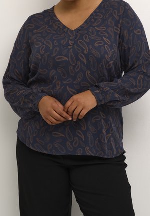 Persoon die een marineblauwe blouse met lange mouwen draagt met bruine paisley-patronen en een zwarte broek, handen zachtjes in elkaar gevouwen voor zich.