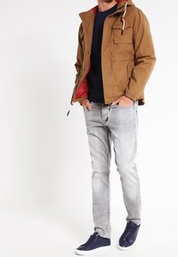 Veste marron avec un intérieur rouge, dotée d'une capuche et d'une poche avant. Associée à un jean gris clair et des baskets bleu foncé.