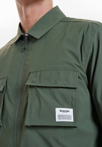 Zielono-oliwkowa kurtka na zamek wykonana z lekkiego materiału, z dwoma kieszeniami na piersi, szpiczastym kołnierzem oraz metką z logo na lewej kieszeni.