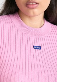 Różowy sweter prążkowany z okrągłym dekoltem i niebieskim haftowanym logo "HUGO" z przodu. Miękka tekstura, dopasowany krój.
