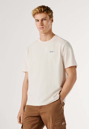 Joven con cabello rubio corto que lleva una camiseta lisa de color blanco hueso y pantalones cargo marrones, posando con una mano en el bolsillo.