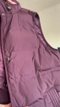 Burgundska prsluk bez rukava s glatkom završnom obradom, koja ima prednji zip zatvarač, kopče na press dugmad i aksentu horizontalnog šivenja.