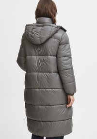 Lang, dunjakke i dempet grå med hette, med en kviltet design og elastiske mansjetter. Glatt, lett strukturert overflate.