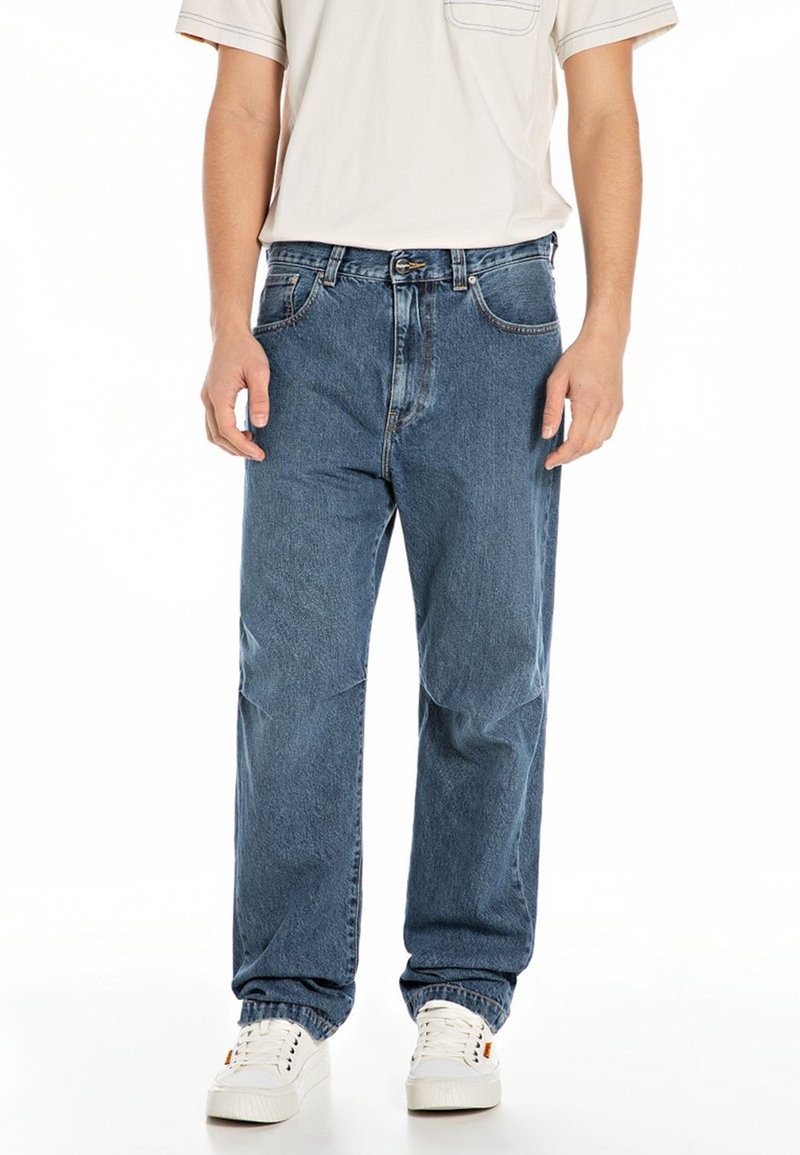Blue denim jeans with a relaxed fit, featuring front pockets, a zip fly, and visible stitching details. Paired with white sneakers.