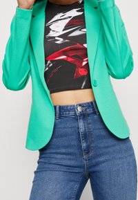 Osoba w miętowej marynarce nałożonej na czarny i czerwony crop top z grafiką, w połączeniu z niebieskimi spodniami typu high-waisted.