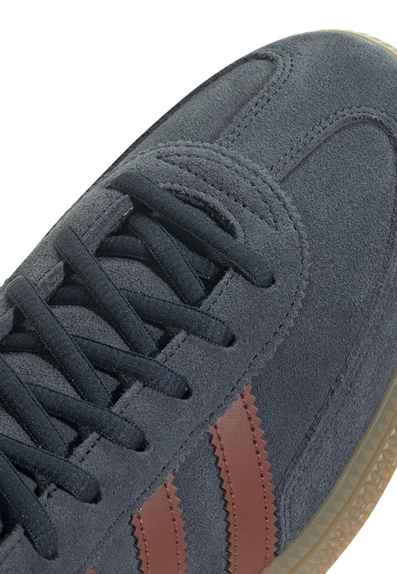 Szare zamszowe sneakersy z ciemnymi sznurowadłami, posiadające brązowe ząbkowane paski po bokach oraz teksturowaną gumową podeszwę.