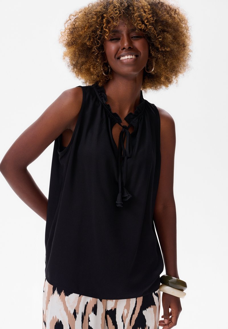 Czarny bezrękawnik z marszczeniem przy dekolcie i detalem w postaci wiązania. Gładki materiał. Modelka nosząca wzorzyste, szerokie spodnie i masywne bransoletki.