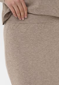 Mano descansando sobre la cinturilla de unos pantalones de punto beige combinados con una parte superior de punto beige, mostrando una tela suave y texturizada.