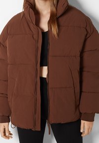 Giubbotto puffo marrone con colletto alto, chiusura con zip e polsini elasticizzati. Presenta trapuntatura orizzontale e tasche laterali. Texture liscia.