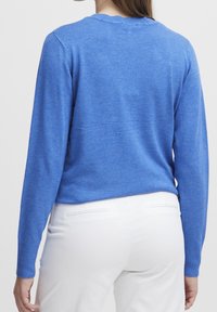 Femme portant un pull bleu à manches longues et un pantalon blanc, vue de dos sur un fond clair uni.