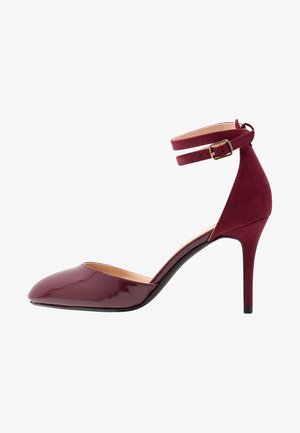 Chaussure à talon haut bordeaux avec un bout pointu, devant en cuir verni et une sangle de cheville en daim avec un détail de boucle en or.