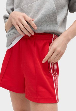 Person iført røde shorts med hvide striber, der peger på en lynlåslomme i siden, iført en grå sweatshirt og en hvid undertrøje.