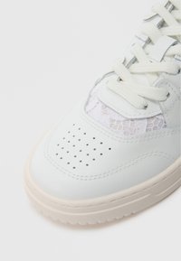 Zapatilla blanca de cuero con un panel de cordones texturizado, diseño perforado en la punta y suela de goma color crema con detalle de costura.