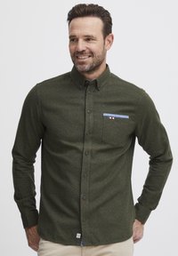 Grønn knapped shirt med en subtil tekstur. Har en brystlomme med blå akcent og logo-tag på nederste kant. Lange ermer.