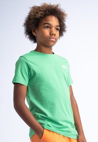 Zelené bavlněné tričko s krátkým rukávem, kulatým výstřihem a malým bílým logem na hrudi. Nosené s oranžovými šortkami.