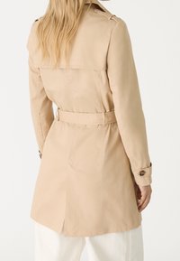 Trench beige con cintura ceñida, mangas largas, hombreras y una abertura en la parte trasera para facilitar el movimiento. Textura de tela suave.