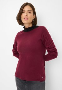 Pullover in maglia bordeaux con scollo rotondo, maniche lunghe e orlo a costine. Presenta una maglia nera a collo alto sovrapposta. Texture morbida.
