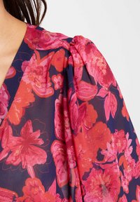 Blusa floral con un vibrante estampado de flores en tonos rosa y rojo sobre un fondo azul marino, con mangas abullonadas y un tejido suave y semi-translúcido.