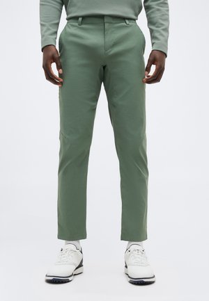 Άνδρας που φοράει στενά παντελόνια ανοιχτού πράσινου χρώματος, λευκές κάλτσες και λευκά παπούτσια με κορδόνια, στέκεται μπροστά σε απλό φόντο.