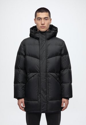 Un homme porte un long manteau matelassé noir à capuche, sur un fond clair uni.