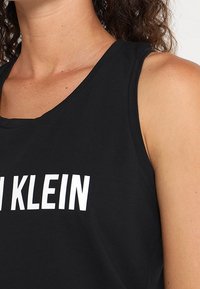 Sort tank top lavet af bomuld med brede ærmegab og rund halsudskæring, fremhævet af store hvide bogstaver, der står "CALVIN KLEIN" på forsiden.