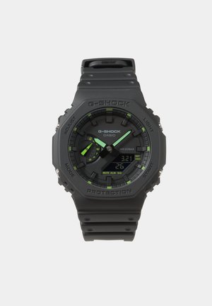 Juodas G-Shock laikrodis su skaitmeniniu ekranu, žaliu akcentu, apvaliu ciferblatu ir tekstūruota guma juosta. Funkcijos apima vandens atsparumą ir kelis nustatymus.