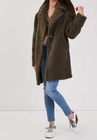 Manteau oversize en fausse fourrure vert olive avec col, deux grands boutons, poches latérales et surface texturée, porté par-dessus un haut à motifs et un jean skinny.