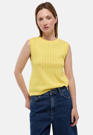 Femme portant un haut tricoté jaune sans manches et un jean bleu, debout avec une main dans le dos et une expression neutre.
