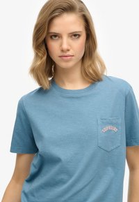 Blauwe casual T-shirt gemaakt van zachte stof, met een borstzak waarin "SUPERDRY" in het roze is geborduurd, ronde halslijn en korte mouwen.