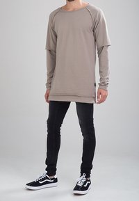 Camisa de manga larga beige con escote redondo y bordes sin terminar, combinada con jeans ajustados negros y zapatillas negras con detalles blancos.