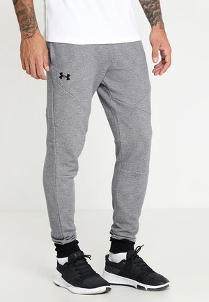 Mężczyzna w szarych dresach Under Armour, czarno-białych butach sportowych oraz białej koszulce, stojący na jednolitym tle.