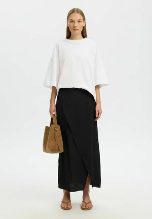 Kvinne med rett blondt hår iført en løs hvit topp, svart omslagskjørt, brune sandaler og bærer en brun semsket skinnveske.