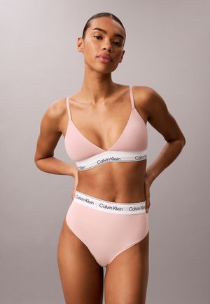 Kvinde iført lyserød Calvin Klein bralette og højtaljede trusser med hvide elastiske bånd med logo, poserer mod en ensfarvet grå baggrund.