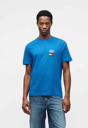 Camiseta de algodón azul con mangas cortas, que presenta un pequeño logo bordado y un parche rectangular en rojo, blanco y azul. Cuello redondo clásico.
