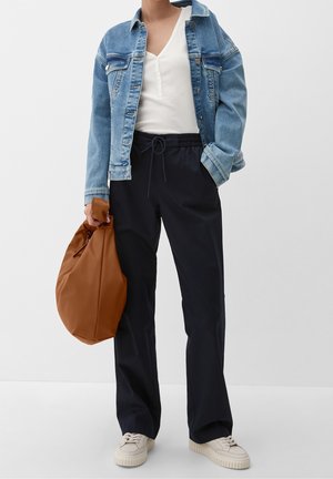 Personne portant une veste en denim bleu, un t-shirt henley blanc, un pantalon noir à cordon de serrage, des baskets blanches, tenant un grand sac marron à la main droite.