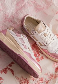Baskets blanches et roses avec des accents en daim beige et des languettes marquées sur un fond en papier rose et blanc à motif floral.