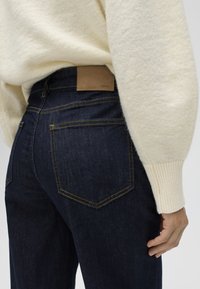 Close-up van een persoon die donkerblauwe spijkerbroek draagt met gele stiksels en een beige gebreide trui, waarbij de achterzak en het leren label op de tailleband zichtbaar zijn.