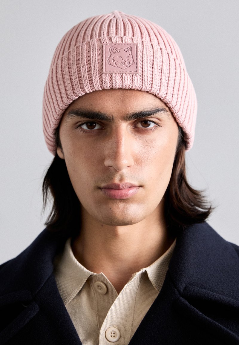 Bonnet en tricot côtelé rose clair avec un tissu texturé et une étiquette carrée avec un design d'animal stylisé à l'avant.