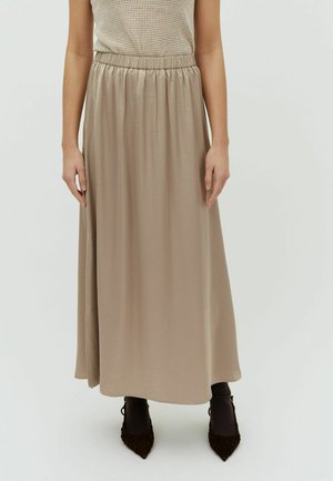 Beżowa długa spódnica z elastycznym pasem w talii, zestawiona z brązowymi butami w szpic i ciemnymi skarpetkami, modelka pokazana od środka tułowia do stóp na jednolitym tle.