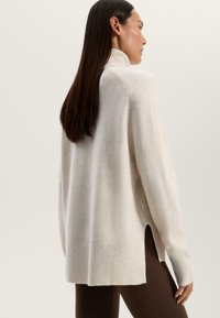 Maglione a collo alto di colore crema realizzato in un tessuto morbido e testurizzato, con spacchi laterali e orlo a coste, abbinato a leggings scuri.