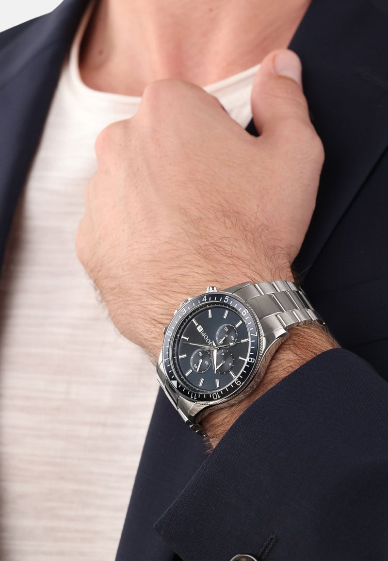 Zilveren roestvrijstalen polshorloge met een zwarte wijzerplaat, drie subdials en een metalen armband. Draagt aan een hand tegen een donkere jas.