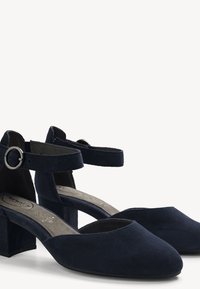 Chaussures en daim bleu marine à talons blocs, bouts pointus et brides de cheville avec boucles rondes argentées sur fond blanc.