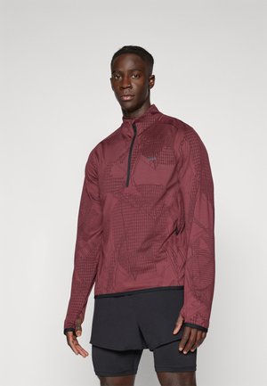 Burgunderfarbener Zip-Kragen-Jacke mit geometrischem Muster, die eine strukturierte Oberfläche und ein tailliertes Design aufweist, kombiniert mit schwarzen Shorts.