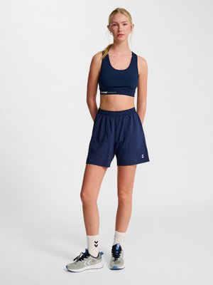 Kobieta stojąca z rękami za plecami, ubrana w granatowy sportowy biustonosz, granatowe sportowe szorty, białe skarpetki i szare buty do biegania na jednolitym tle.