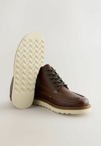 Brune lædersko med moc-toe design, kontrasterende syninger og en let cremefarvet ydersål med zigzag mønster.